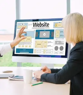 Two people collaborate on a desktop monitor showing a detailed website wireframe layout