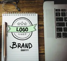 Notebook showing a hand-drawn emblem for a LOGO and the words BRAND IDENTITY next to a pen and laptop.